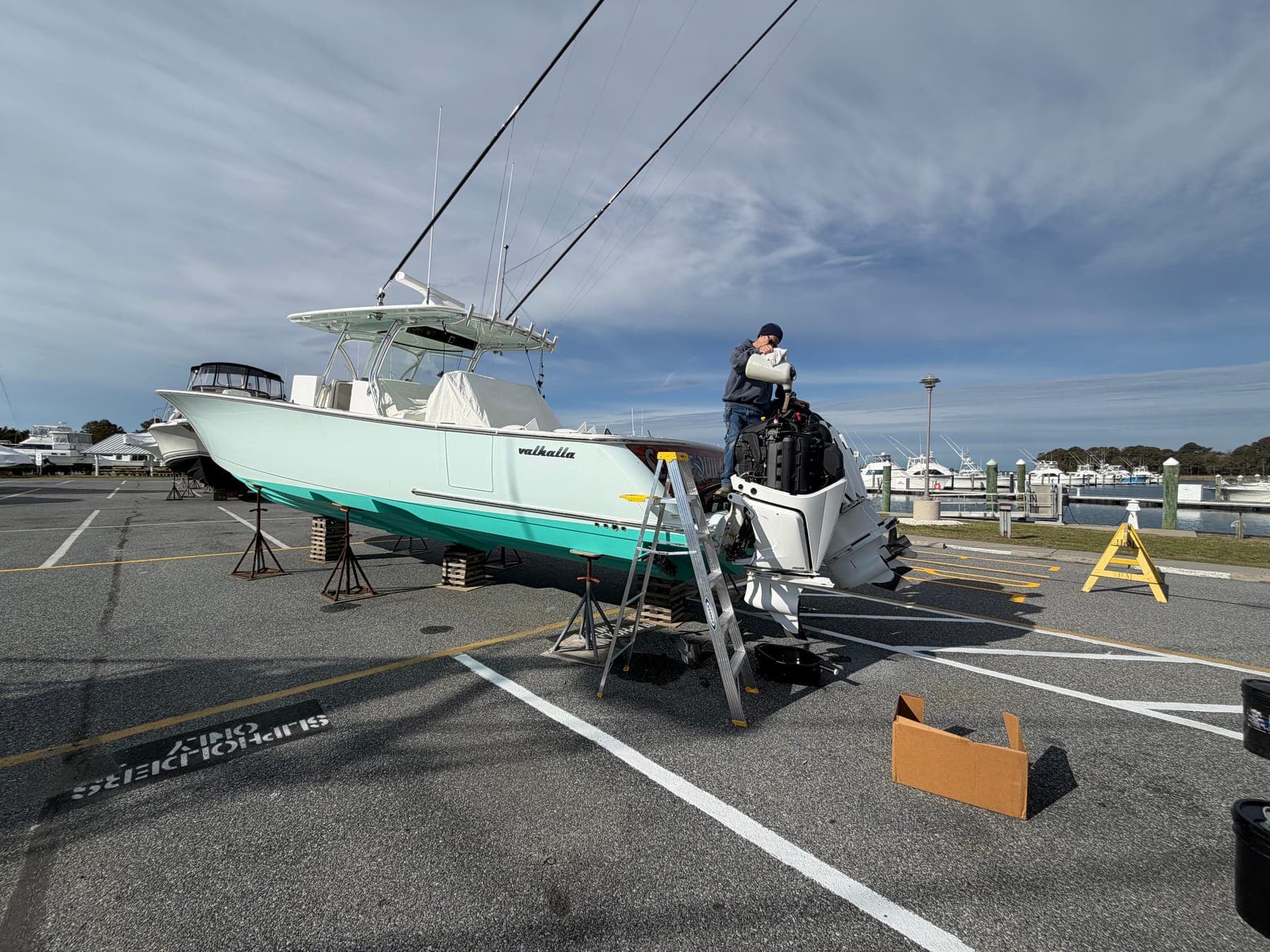 A Delmarva Service Center technician standing at the transom of a large seafoam-green center console up on stands at a marina, pouring oil into an outboard with the cowling removed